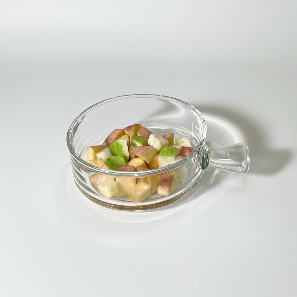 Two clear ribbed glass bowls with handles are shown on a white tabletop. One bowl sits on the table holding mixed fruit pieces, and the other is held by a person's hand, also containing fruit.